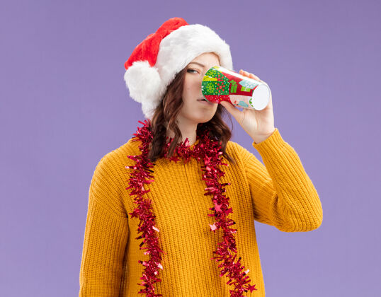 年轻自信的年轻斯拉夫女孩戴着圣诞帽 脖子上戴着花环 喝着纸杯里的酒 背景是紫色的 有复印空间周围花环斯拉夫人