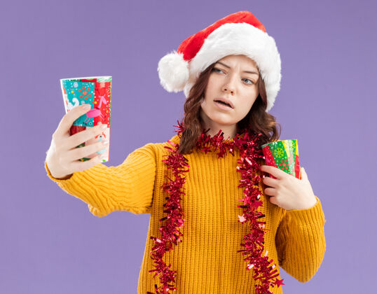 花环自信的年轻斯拉夫女孩 戴着圣诞帽 脖子上戴着花环 拿着纸杯 看着紫色背景上的纸杯 还有复制空间周围圣诞老人自信