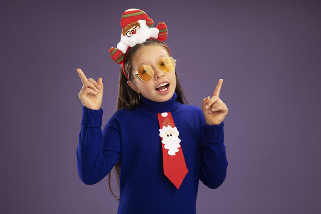 领带小女孩穿着蓝色高领毛衣 系着红色领带 头上戴着有趣的圣诞戒指 看着镜头 快乐而欢快地用食指站在紫色的背景上站立圣诞节小