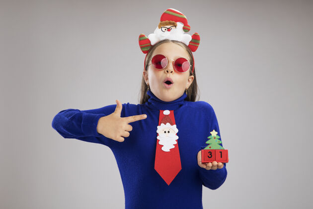 日期穿蓝色高领毛衣的小女孩头上戴着有趣的圣诞戒指 手里拿着玩具立方体 新年快乐 看起来很惊讶 用食指指着站在白色背景上的立方体站着立方体圣诞节