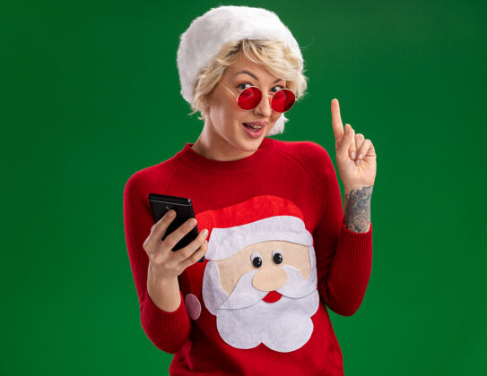 印象深刻给人印象深刻的年轻金发女人戴着圣诞帽和圣诞老人的圣诞毛衣戴着眼镜拿着手机看着并指着隔离在绿色墙上的复制空间女人手机圣诞老人