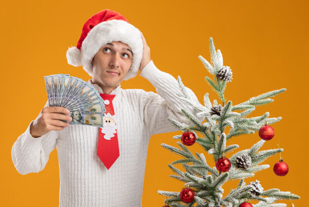 帽子困惑的年轻帅哥戴着圣诞帽 打着圣诞老人的领带 站在装饰好的圣诞树旁 手里拿着钱 手放在头上 看着橙色墙上孤立的一面困惑穿着头
