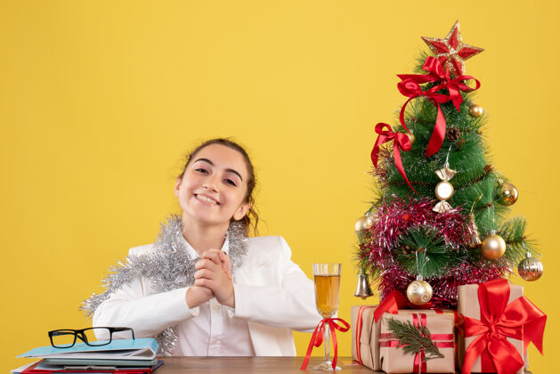 人正面图：女医生坐在桌子后面 在黄色背景下微笑着 手里拿着圣诞树和礼品盒微笑办公室医生