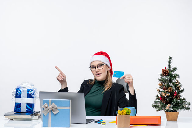 圣诞节一个戴着圣诞老人帽子戴着眼镜的漂亮女人坐在桌旁 手里拿着圣诞礼物和银行卡 在办公室股票的形象眼镜行政人员穿着