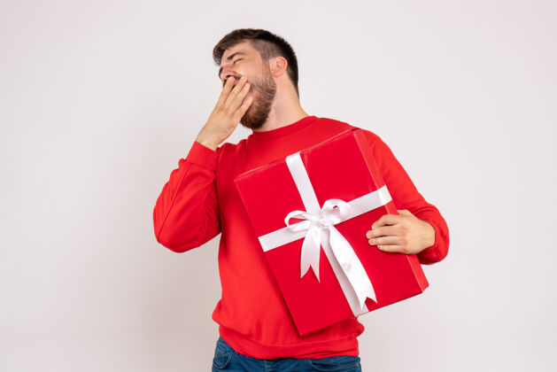 年轻身穿红衫的年轻人手持圣诞礼物 在白墙上打哈欠的正面图男性礼物抱着