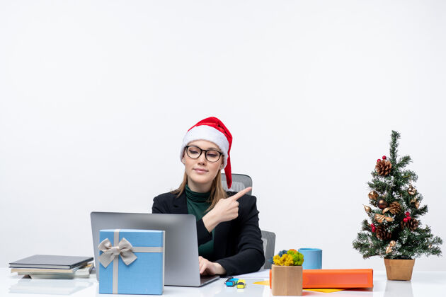 礼物戴着圣诞老人帽的女商人坐在桌子旁 桌上放着圣诞树和礼物工作场所笔记本电脑商务