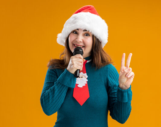 胜利微笑的成年白人妇女 戴着圣诞帽 打着圣诞领带 拿着麦克风 打着胜利的手势 橙色背景上有复制空间手势快乐麦克风