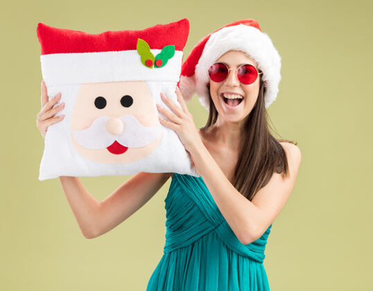 女孩快乐的年轻白人女孩戴着太阳眼镜 戴着圣诞帽 抱着圣诞枕头 背景是橄榄绿 有复印空间新年橄榄举行