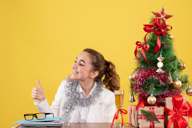 快乐前视图：女医生坐在桌子后面 拿着圣诞礼物和黄色背景上的树新郎人礼物