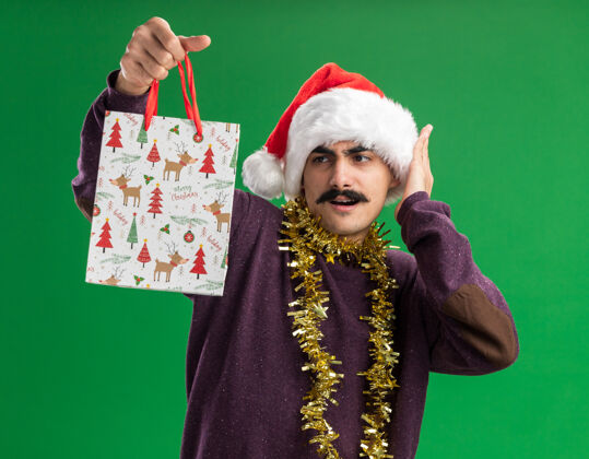 圣诞快乐年轻的胡子男人戴着圣诞老人帽子 脖子上挂着金属丝 手里拿着圣诞礼物的纸袋 站在绿色的背景下 困惑而惊讶地看着它圣诞老人围着惊讶