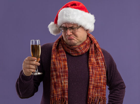香槟困惑的成年男子戴着眼镜 戴着圣诞帽 脖子上围着围巾 手里拿着一杯孤立在紫色背景上的香槟圣诞快乐困惑成人