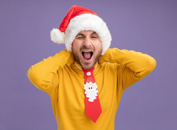 相机自信的白人年轻人戴着圣诞帽 打着领带 双手放在脖子后面 看着摄像机眨着眼睛 张着嘴 与紫色背景隔离脖子帽子手