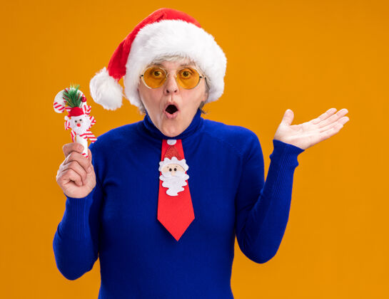 打开一位戴着太阳眼镜 戴着圣诞帽 打着圣诞领带 手持糖果手杖 手一直张开的老妇人惊讶地站在橙色背景上 留着复印空间领带老人手杖
