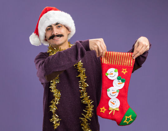 圣诞节年轻的胡子男人戴着圣诞老人帽 脖子上挂着金属丝 手里拿着圣诞长袜 看着摄像机 脸上洋溢着幸福的笑容 站在紫色的背景下 欢快地笑着长袜站着金属丝