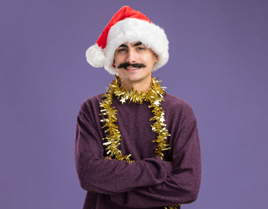 帽子快乐的小胡子男人戴着圣诞老人的帽子 脖子上戴着金属丝 站在紫色的背景下 面带微笑地看着相机圣诞节金属片脸
