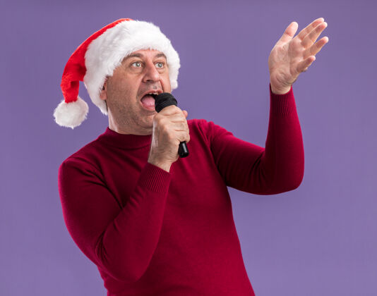 举行中年男子戴着圣诞老人的帽子拿着麦克风站在紫色的背景上快乐而兴奋地唱歌年龄兴奋唱歌