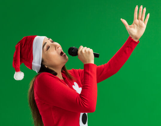 圣诞老人戴着圣诞老人帽 穿着红色毛衣的年轻女子手持麦克风高高兴兴地唱着歌 她举起手臂站在绿色的背景上站着唱歌兴奋