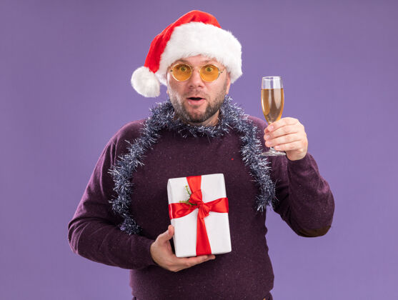 花环令人印象深刻的中年男子戴着圣诞帽 脖子上戴着金箔花环 戴着眼镜 手里拿着礼包 紫色墙上隔着一杯香槟包装中年男人