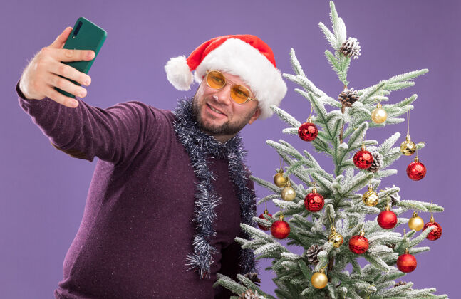 脖子微笑的中年男子戴着圣诞帽 脖子上戴着金箔花环 戴着眼镜 站在装饰好的圣诞树旁 独自在紫色的墙上自拍穿着金属片附近