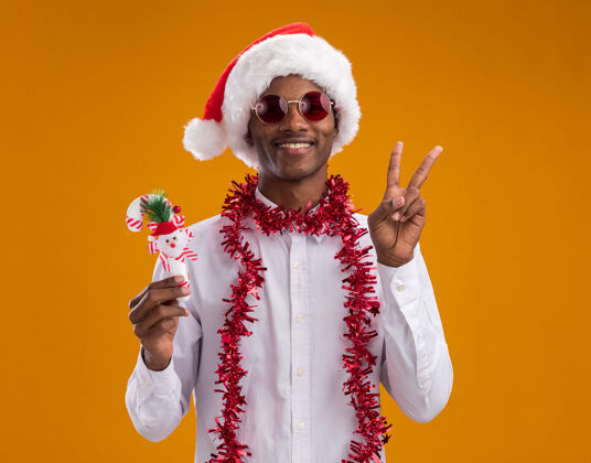 帽子微笑着的年轻的非洲裔美国人戴着圣诞帽 戴着眼镜 脖子上戴着金箔花环 手里拿着糖果手杖做着和平的手势 隔离在橙色的墙上男人圣诞节圣诞老人