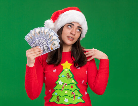 年轻戴着圣诞帽的恼怒的白人女孩拿着钱看着在绿色背景上与世隔绝的一面女孩烦恼持有