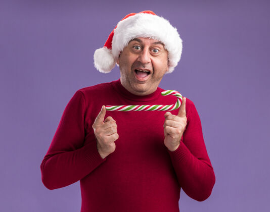 圣诞节中年男子戴着圣诞老人的帽子拿着糖果手杖看着相机快乐而兴奋地站在紫色的背景上举行中间兴奋