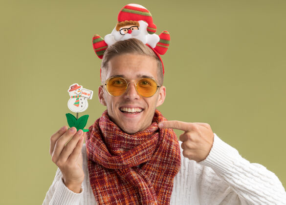 雪人快乐的年轻帅哥戴着圣诞老人的头带和围巾拿着和指着橄榄绿墙上孤立的雪人玩具围巾圣诞老人圣诞节