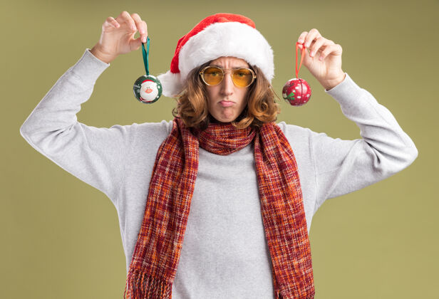 周围沮丧的年轻人戴着圣诞老人的帽子 戴着黄色的眼镜 脖子上围着暖和的围巾 手里拿着圣诞球 站在绿色的背景下 带着悲伤的表情看着摄像机心烦球悲伤