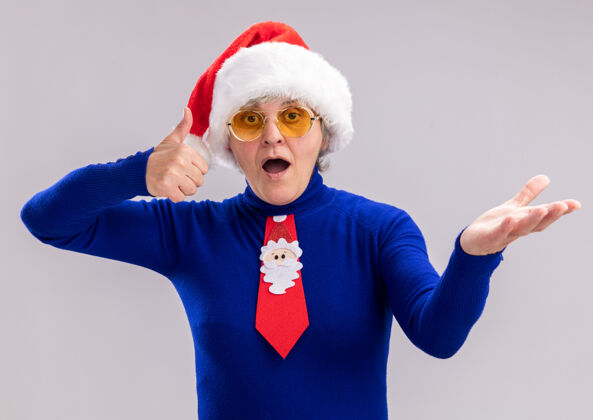 印象深刻一位戴着太阳眼镜 戴着圣诞帽 系着圣诞领带的老妇人 竖起大拇指 在白色背景上 双手张开 并留有复印空间圣诞老人帽子手
