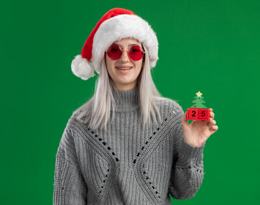立方体年轻的金发女郎穿着冬季毛衣 戴着圣诞帽 戴着红眼镜 手里拿着玩具立方体 手里拿着圣诞节的日期 看着相机 站在绿色的背景下开心而积极地微笑着圣诞节毛衣微笑