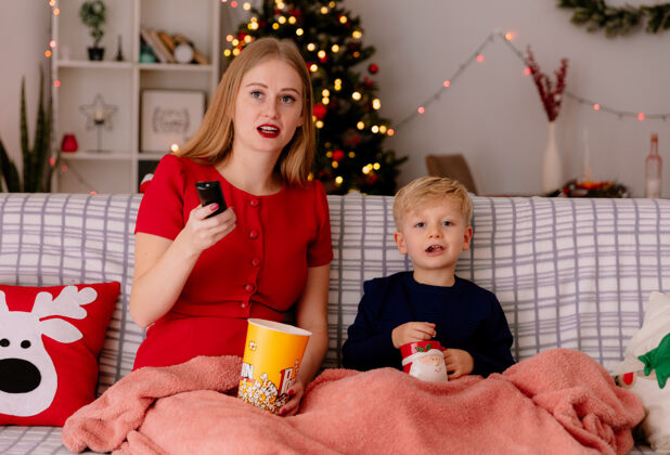沙发快乐的妈妈穿着红色的裙子 带着她的孩子坐在沙发上 毯子下面放着一桶爆米花 在装饰好的房间里一起看电视 背景是圣诞树房间圣诞树衣服