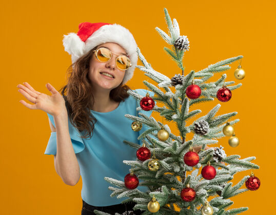 圣诞身着蓝色上衣 戴着圣诞帽 戴着黄色眼镜装饰圣诞树的快乐而积极的年轻女子看着镜头挥舞着手臂站在橙色背景上帽子装饰圣诞老人