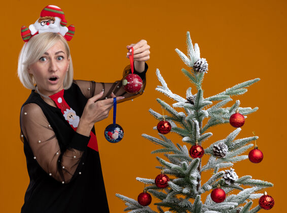 期待惊讶的中年金发女人戴着圣诞老人的头带和领带站在侧面图装饰圣诞树附近拿着圣诞饰品看着橙色背景上孤立的相机个人资料举行视图