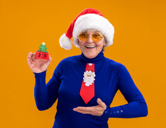 圣诞戴着太阳眼镜 戴着圣诞帽 系着圣诞领带的快乐的老妇人拿着圣诞树的装饰物 在橙色的背景上与复制空间隔离开来拿着指着圣诞老人