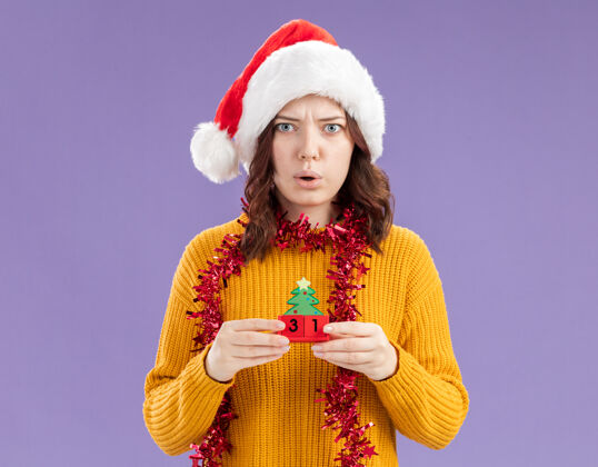 帽子令人印象深刻的年轻斯拉夫女孩与圣诞帽和花环围着脖子举行圣诞树装饰孤立的紫色背景与复制空间周围装饰品年轻