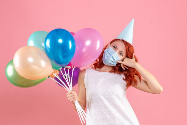 年轻女性前视图中的年轻女子拿着五颜六色的气球在无菌面具上粉红色的墙壁女性庆祝气球