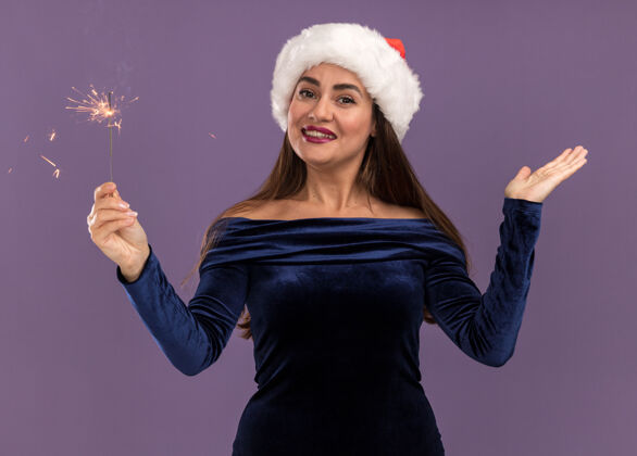 闪闪发光微笑着的年轻漂亮的女孩穿着蓝色的裙子 戴着圣诞帽 手里拿着闪闪发光的东西 在紫色的背景上孤立着手女孩微笑帽子