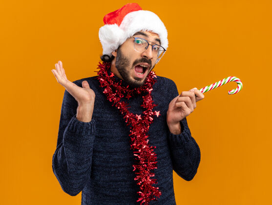 抱着惊讶的年轻帅哥戴着圣诞帽 脖子上戴着花环 手里拿着圣诞糖果 手被隔离在橙色的背景上惊讶蔓延糖果