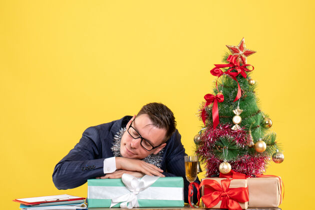 礼物昏昏欲睡的商人坐在圣诞树旁的桌子前 黄色的礼物坐着桌子新郎