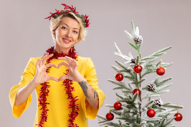 树微笑着的金发女郎头戴圣诞花环 脖子上戴着金属丝花环 站在装饰好的圣诞树旁 看着摄像机 在白色背景上做着心形标志年轻微笑头