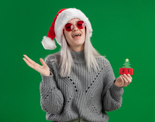 立方体年轻的金发女郎穿着冬衣 戴着圣诞帽 戴着红眼镜 手里拿着玩具立方体 在绿色的背景下欢快地站着 带着快乐的新年约会 快乐而积极的微笑年轻年帽子