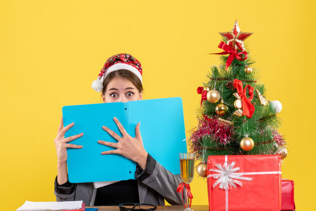 女孩前视图震惊的女孩与圣诞帽坐在桌上覆盖着她的脸与文件文件夹圣诞树和礼物鸡尾酒坐着帽子文件
