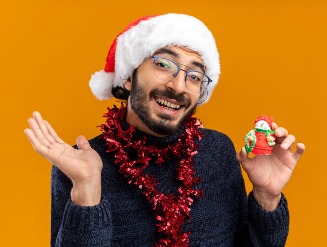 小伙子微笑的年轻帅哥戴着圣诞帽 脖子上戴着花环 手里拿着玩具 手被隔离在橙色的背景上抱着微笑帽子