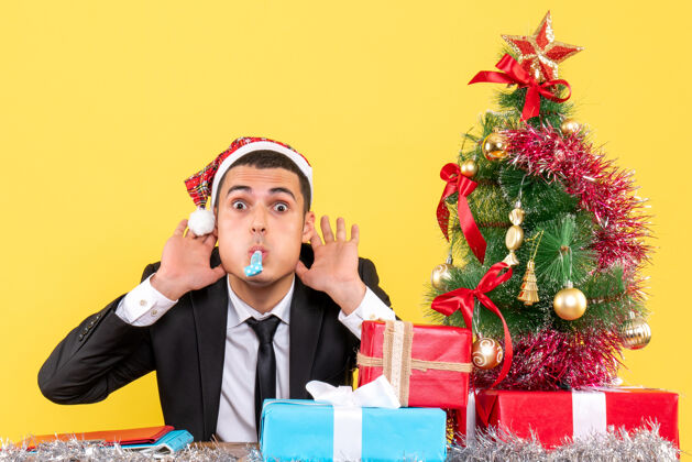 帽子正面图：戴着圣诞帽的年轻人坐在桌子旁 用噪音制造圣诞树和礼物噪音制造者坐着西装