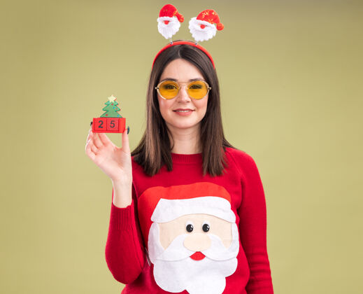 漂亮高兴的年轻漂亮的白人女孩穿着圣诞老人毛衣和戴着眼镜的头带拿着圣诞树玩具和日期看着相机隔离在橄榄绿的背景毛衣圣诞树橄榄