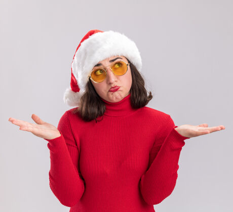 伸展穿着红色毛衣 戴着圣诞帽 戴着眼镜的年轻女孩看起来很困惑 双臂伸向两边 站在白色的背景上年轻侧面站立