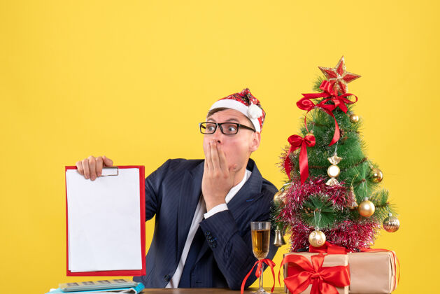惊奇的人前视图惊讶的人拿着剪贴板坐在圣诞树旁的桌子上 黄色的墙上摆着礼物桌子人办公室
