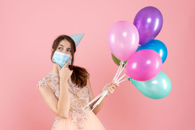 医疗戴着派对帽和医用面具的快乐女孩手持五颜六色的气球 手放在粉红色的下巴上面具女孩下巴