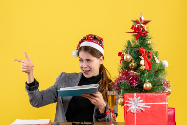 文件夹正面图年轻女孩戴着圣诞帽坐在桌旁拿着文件夹圣诞树和鸡尾酒礼物鸡尾酒树礼物