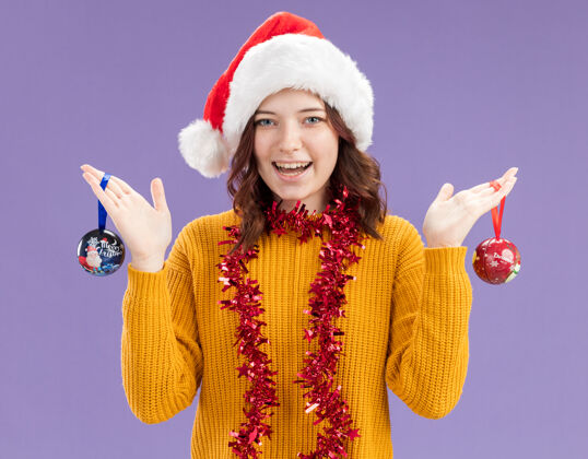 脖子快乐的年轻斯拉夫女孩 戴着圣诞帽 脖子上戴着花环 手拿玻璃球饰品 紫色背景上有复制空间球快乐圣诞老人